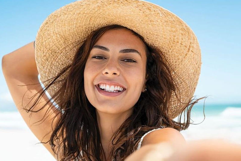 Picture of a on the beach, happy with her plastic surgery at Cancun MedVentures.  The woman is wearing tan sun hat and sitting on a Cancun beach.