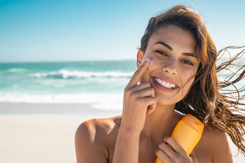 Picture of a smiling woman, happy with her nose surgery she had at Cancun MedVentures.  The woman has long brown hair and is standing on a sandy Cancun beach with the ocean in the background.