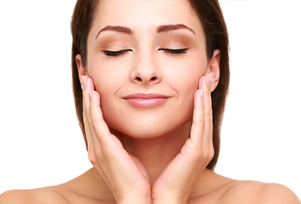 Picture of a woman with long dark brown hair, facing the camera with eyes closed, holding her hands to the side of the cheeks and happy with her perfect face lift with neck lift procedure she had with top plastic surgeons in beautiful Cancun, Mexico.